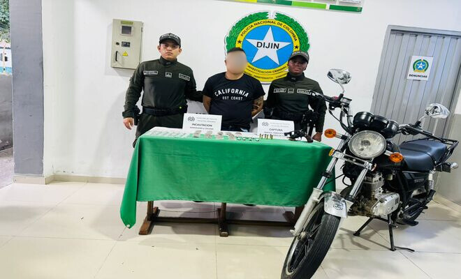 Dos funcionarios de la Policía Nacional presentan a una persona junto a una mesa con elementos incautados y una motocicleta.